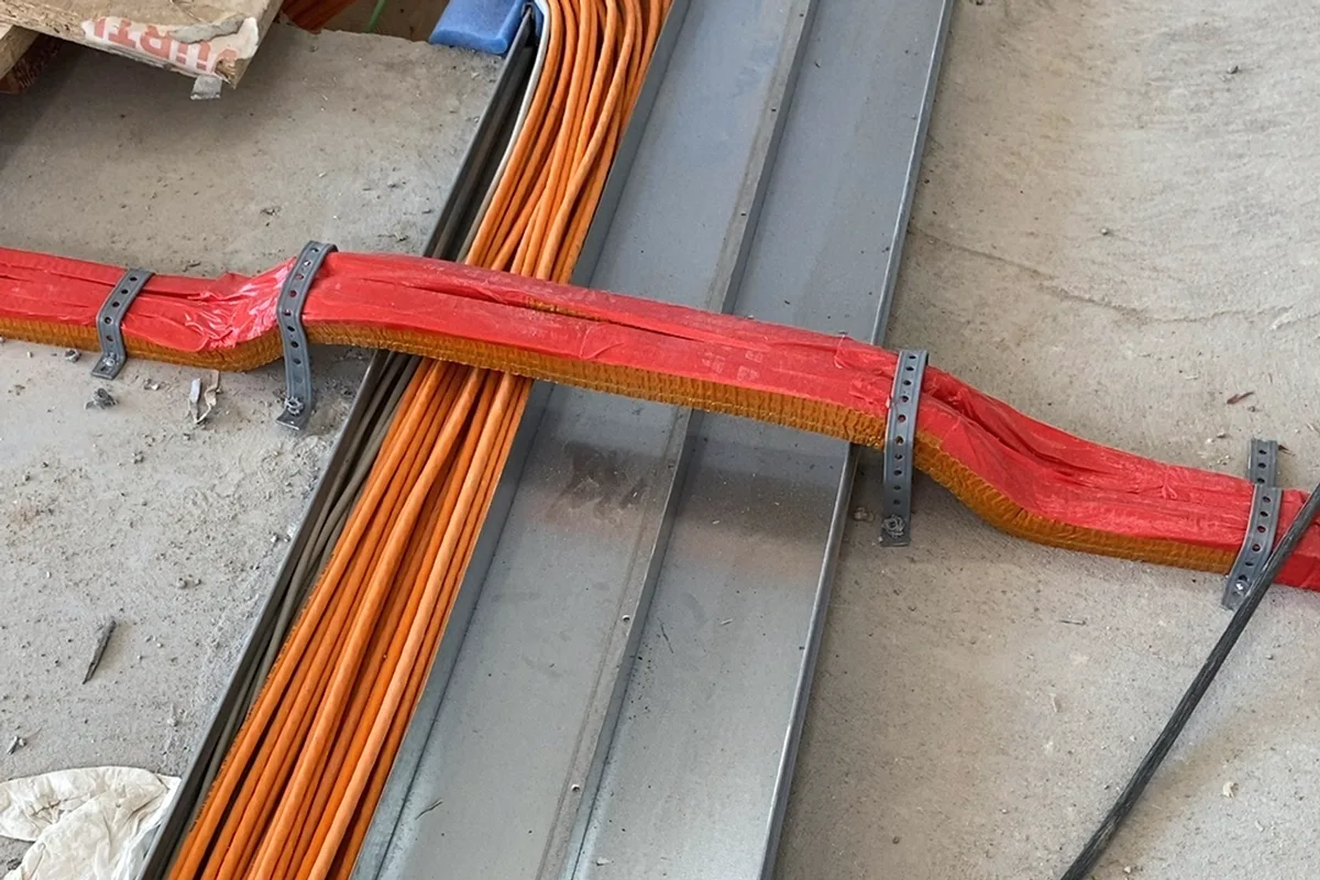 Sottofondo grezzo: un tubo con isolamento rosso incrocia un fascio di cavi arancione e un canale di ventilazione